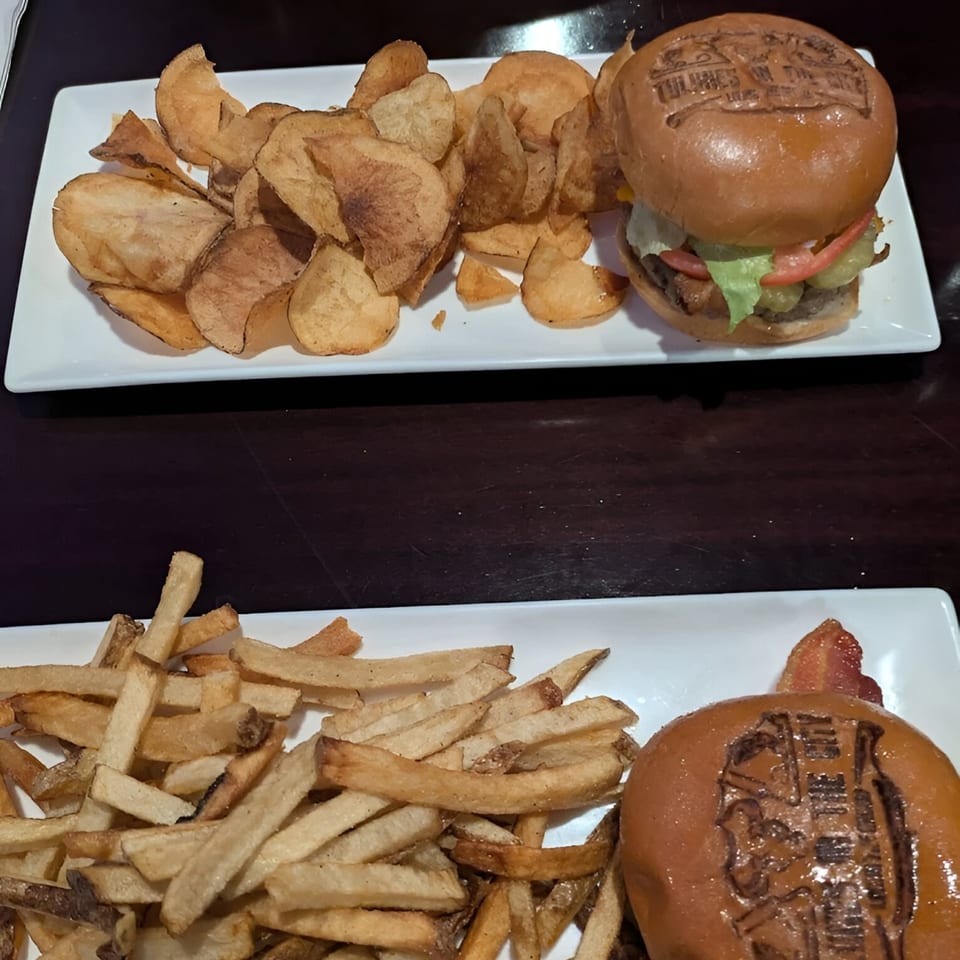 Two Tulunes In The City burgers, one with chips and one with fries, on white plates.