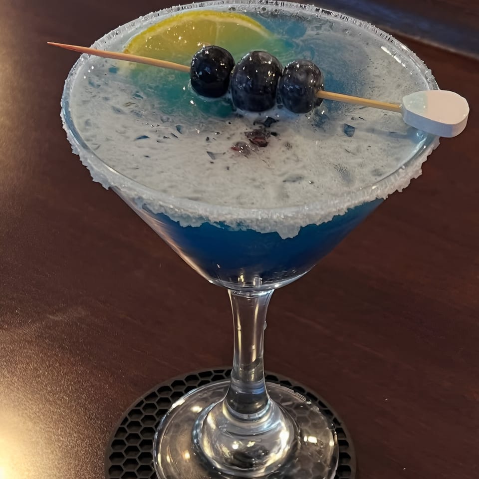 A blue cocktail in a martini glass, garnished with blueberries, a lemon slice, and a sugar rim.