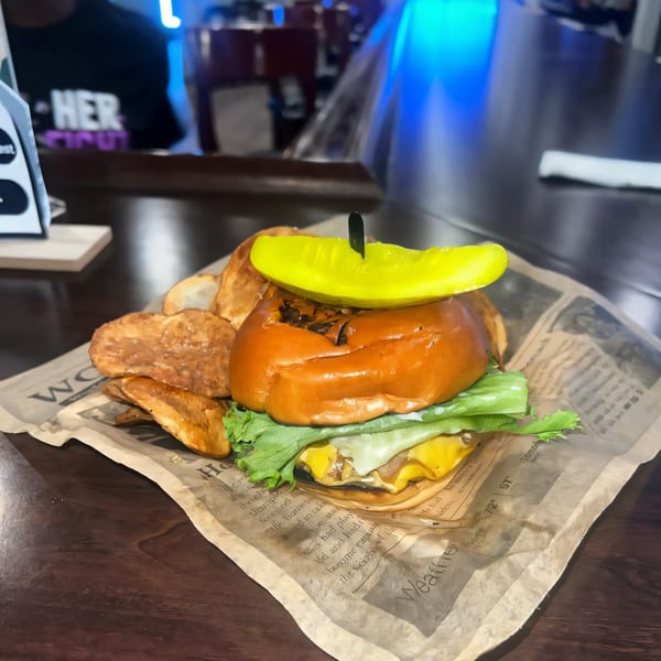 A Tulunes In The City burger with lettuce, cheese, and a pickle, served with potato chips.